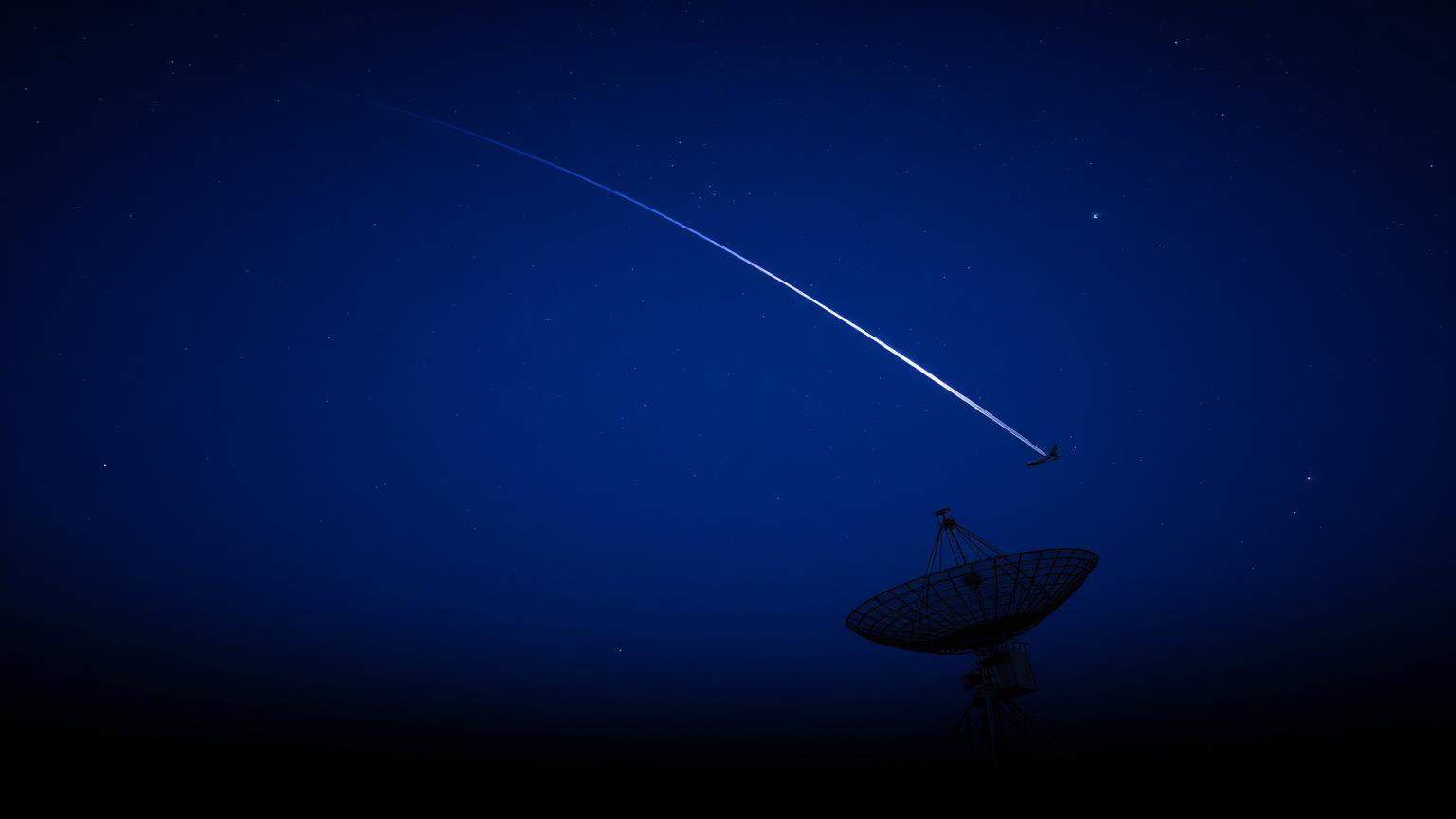 Long-exposure night sky photo of a single aircraft trail above a silhouette radar dish